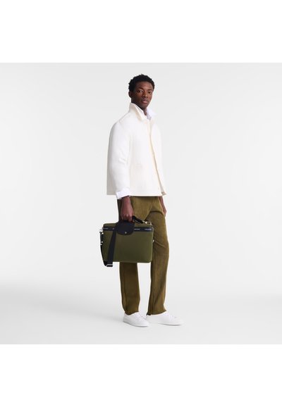 A man holds a green briefcase with a black handle, wearing a white linen jacket, white shirt, and olive-colored pants, with white sneakers.