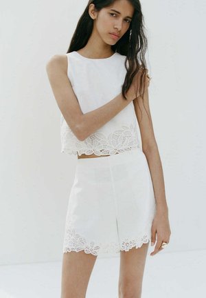 Mujer con cabello largo y oscuro, vestida con una camiseta sin mangas blanca y pantalones cortos a juego con ribete de encaje, de pie frente a un fondo blanco liso.