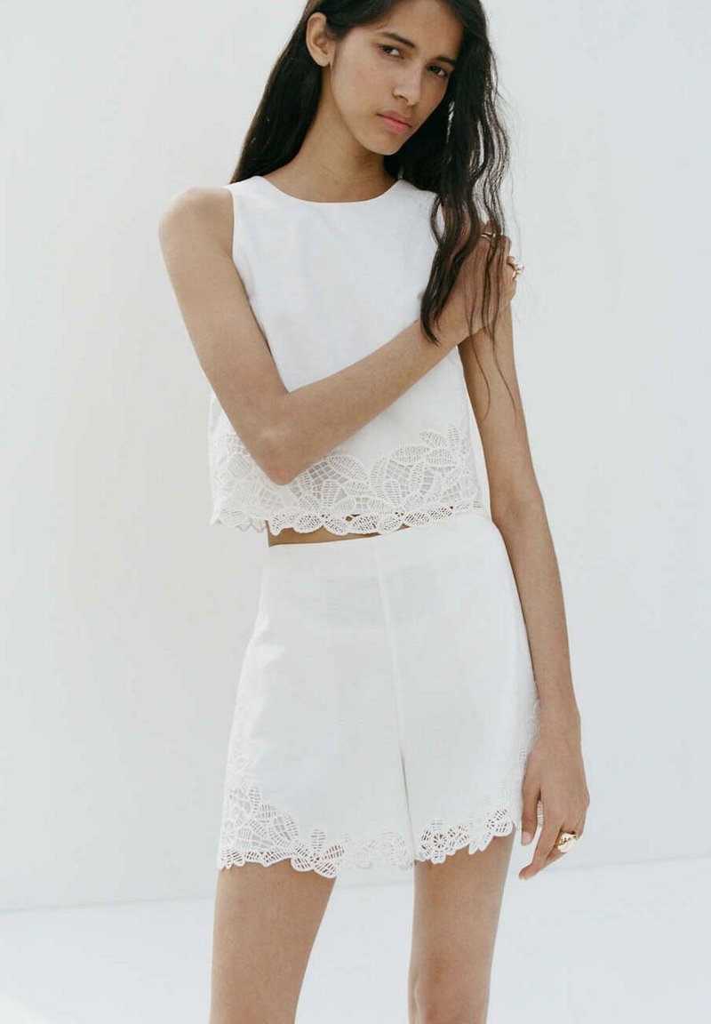 Femme aux longs cheveux foncés portant un haut blanc sans manches et un short assorti avec une bordure en dentelle, debout devant un fond blanc uni.