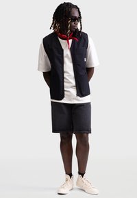 Svart ermeløs vest over en hvit t-skjorte, kombinert med mørke shorts og beige høyhælte sneakers. Har en brystlomme og avkappede ermer.