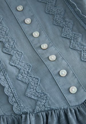 Lichtblauwe katoenen blouse met geborduurde zigzag- en bloemmotieven, zes knoopsluitingen en een geplooide zoomdetail.