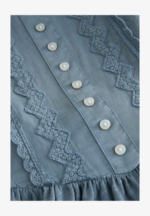 Lichtblauwe katoenen blouse met geborduurde zigzag- en bloemmotieven, zes knoopsluitingen en een geplooide zoomdetail.