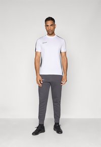 Bela atletska majica s crnimi ramenskimi akcenti in Nike logotipom, v kombinaciji s temno sivo športno hlačami in črnimi športnimi čevlji. Enostaven dizajn.