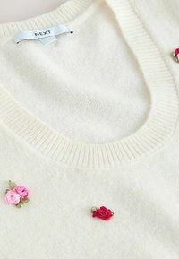 Roomkleurige gebreide trui met een brede ronde halslijn. Voorzien van drie geborduurde bloemenaccenten in roze en rood aan de voorkant. Zachte textuur.