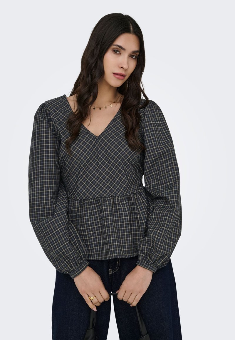 Femme aux longs cheveux foncés portant un chemisier à carreaux sombre à col en V avec des manches longues bouffantes, debout et tenant un sac à main noir.