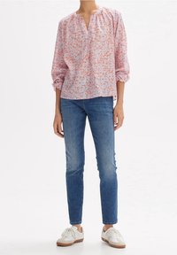 Blouse à motifs floraux dans des tons de rose clair et d'orange, avec un col en V, de longues manches bouffantes et une coupe décontractée. Associée à un jean bleu et des baskets blanches.
