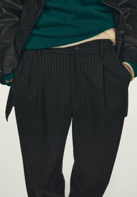 Schwarze Nadelstreifenhose mit Seitentaschen, vorne mit Falten. Der Stoff hat eine dezente Struktur, kombiniert mit einem grünen Pullover darunter.