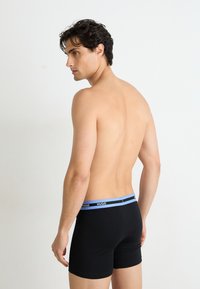 Boxers negros con una cinturilla elástica azul y negra, que presentan el nombre de la marca "HUGO." Modelo con cabello rizado de espaldas.