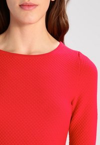 Pull rouge texturé avec un col rond, présentant un motif en losanges et des manches longues, fabriqué à partir d'un tissu extensible et tissé.