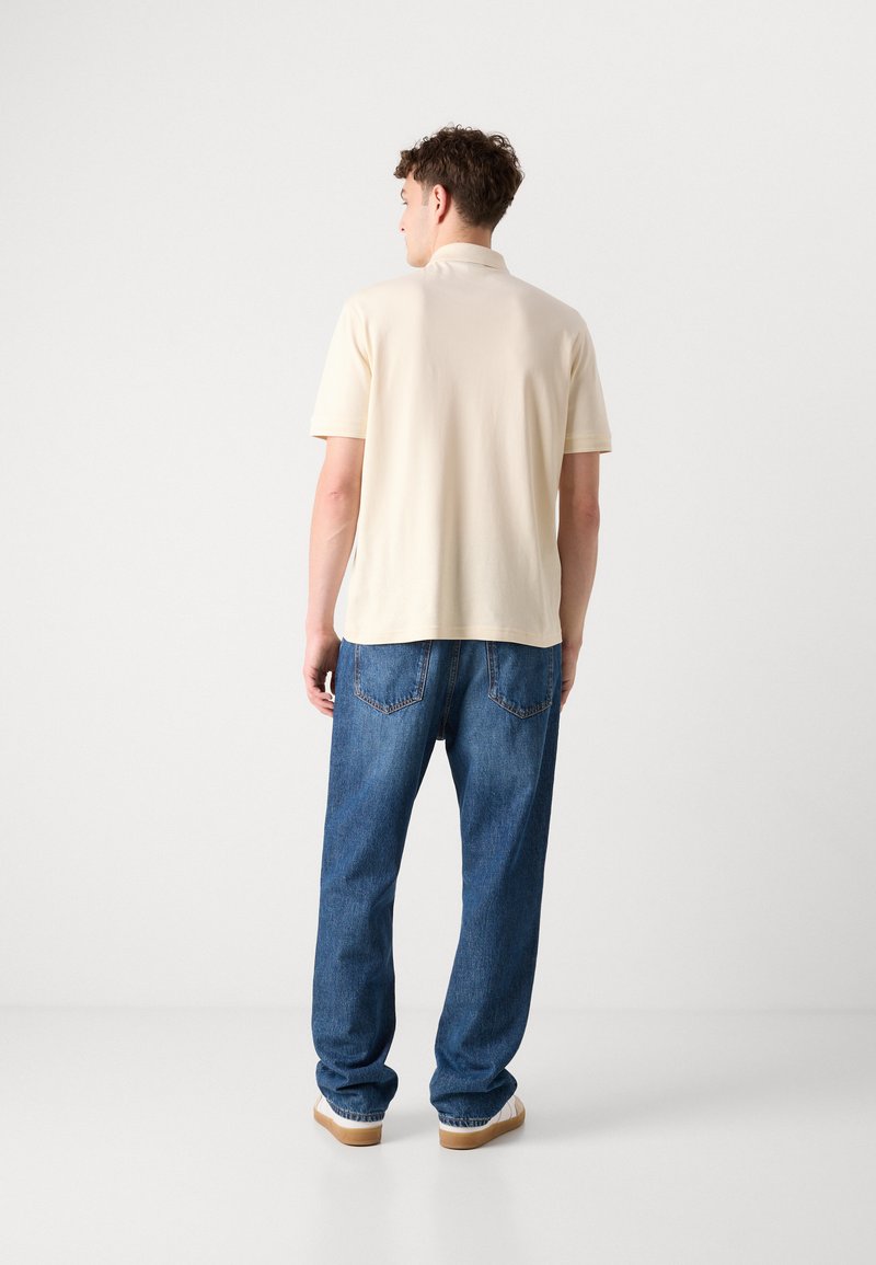 Chemise polo crème et jean bleu ample. La chemise a des manches courtes et un col classique. Le jean a cinq poches et une texture délavée.