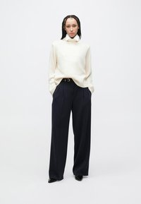 Κρεμ πουλόβερ σε oversize γραμμή με ριγέ λαιμόκοψη, σε συνδυασμό με παντελόνια σε ναυτικό μπλε, ψηλόμεσα και φαρδιά. Λείες υφές και μινιμαλιστικός σχεδιασμός.