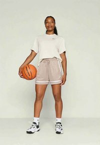 Mujer de pie sosteniendo un balón de baloncesto con la mano derecha, vestida con pantalones cortos deportivos beige, camiseta blanca, calcetines blancos y zapatillas en blanco y negro.