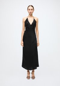 Black halter dress featuring a deep V-neck and sequined bodice, with a smooth flowing skirt and side slit. Model wearing strappy black sandals.