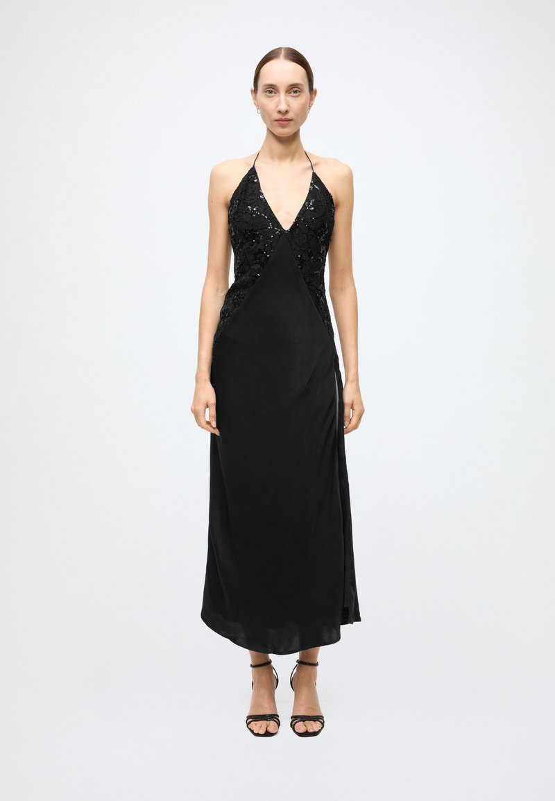 Black halter dress featuring a deep V-neck and sequined bodice, with a smooth flowing skirt and side slit. Model wearing strappy black sandals.
