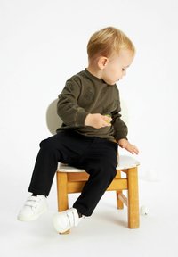 Sudadera verde oliva con estampados de animales en negro, combinada con pantalones negros y zapatos de velcro blancos. Niño pequeño sentado en una silla de madera.