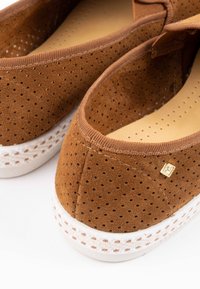 Bruine geperforeerde suede instappers met een witte zool, voorzien van geweven details en een klein gouden logo-accent aan de zijkant.