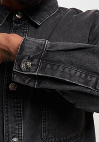 Svart denimjakke med strukturert krage, knappede mansjetter og sølvfarget maskinvare. Stoffet har en glatt, lett teksturert overflate.