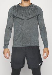 Mann in einem grauen gemusterten Langarm-Sportshirt und schwarzen Nike-Shorts über schwarzen Leggings, stehend mit entspannten Armen.