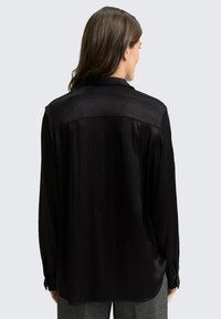 Chemise noire à manches longues en tissu brillant, avec une texture lisse, une coupe droite et des poignets boutonnés. La vue arrière montre un détail de passepoil.