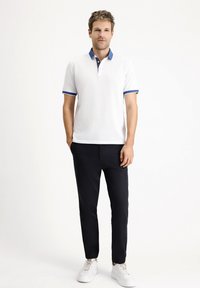 Camiseta polo blanca con cuello y acentos en las mangas azules, combinada con pantalones negros y zapatillas blancas; cuenta con una tapeta de dos botones y una tela texturizada.