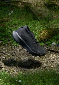 Schwarzer Wanderschuh mit strukturiertem Sohlenprofil und "TERREX"-Markierung, der über felsigem Boden mit grünem Gras und kleinen Blumen schwebt.