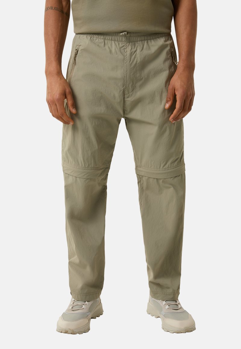 Helle khakifarbene Hose mit einem elastischen Bund, seitlichen Reißverschlusstaschen, geradem Schnitt und subtiler Struktur. Kombiniert mit hellen Sneakers.