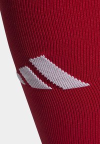 Chaussette rouge montante avec une texture lisse et côtelée, dotée d'un logo stylisé blanc. La chaussette est fabriquée en matériau élastique pour plus d'étirement.