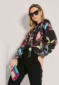 Frau, die eine schwarze Bluse mit Schmetterlingsdruck und Sonnenbrille trägt, eine farbenfrohe gestreifte Clutch hält und vor einer schlichten hellen Wand steht.