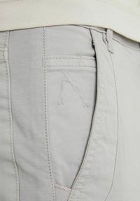 Lichtgrijze broek met een gladde textuur, zijzak en subtiele gestikte logo-detail nabij de zak. Eenvoudig ontwerp met minimale hardware.
