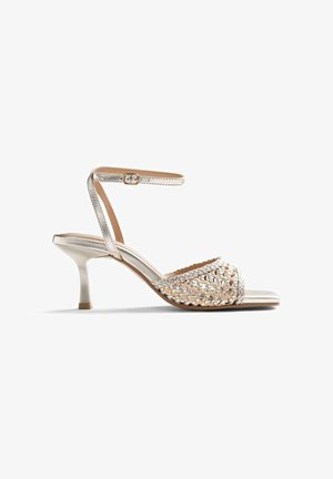 Sandalia de tacón alto plateada con puntera abierta, tira frontal tejida, delgada correa en el tobillo con hebilla y tacón cónico de altura media.