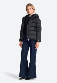 Zwarte pufferjack met capuchon, ritssluiting aan de voorkant en zijzakken, in combinatie met donkere flared jeans en puntige zwarte schoenen.