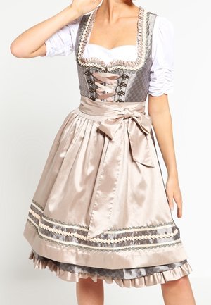 Femme portant une robe traditionnelle dirndl beige et grise avec un corsage à lacets, une blouse blanche et un tablier en satin noué d'un grand nœud à la taille.