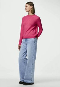 Ženska stoji z rokama v žepih, oblečena v svetlo roza pulover, svetlo modre široke hlače in črne čevlje, obrnjena levo.