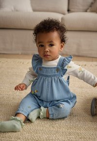 Salopette en denim avec des bretelles volantes, ornée de broderies florales de différentes couleurs. Portée sur une chemise à manches longues blanche et des chaussettes de couleur menthe.