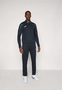 Črna zip-up športna jakna s prilegajočimi hlačami, ki vključuje logotip na prsih in stranske črte. Material se zdi gladek z oprijetim dizajnom.
