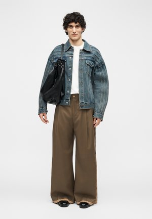 Mladý človek s kučeravými vlasmi oblečený v modrej rifľovej bunde, bielom tričku, širokých hnedých nohaviciach, čiernych topánkach a nesúci veľkú čiernu tašku cez rameno.