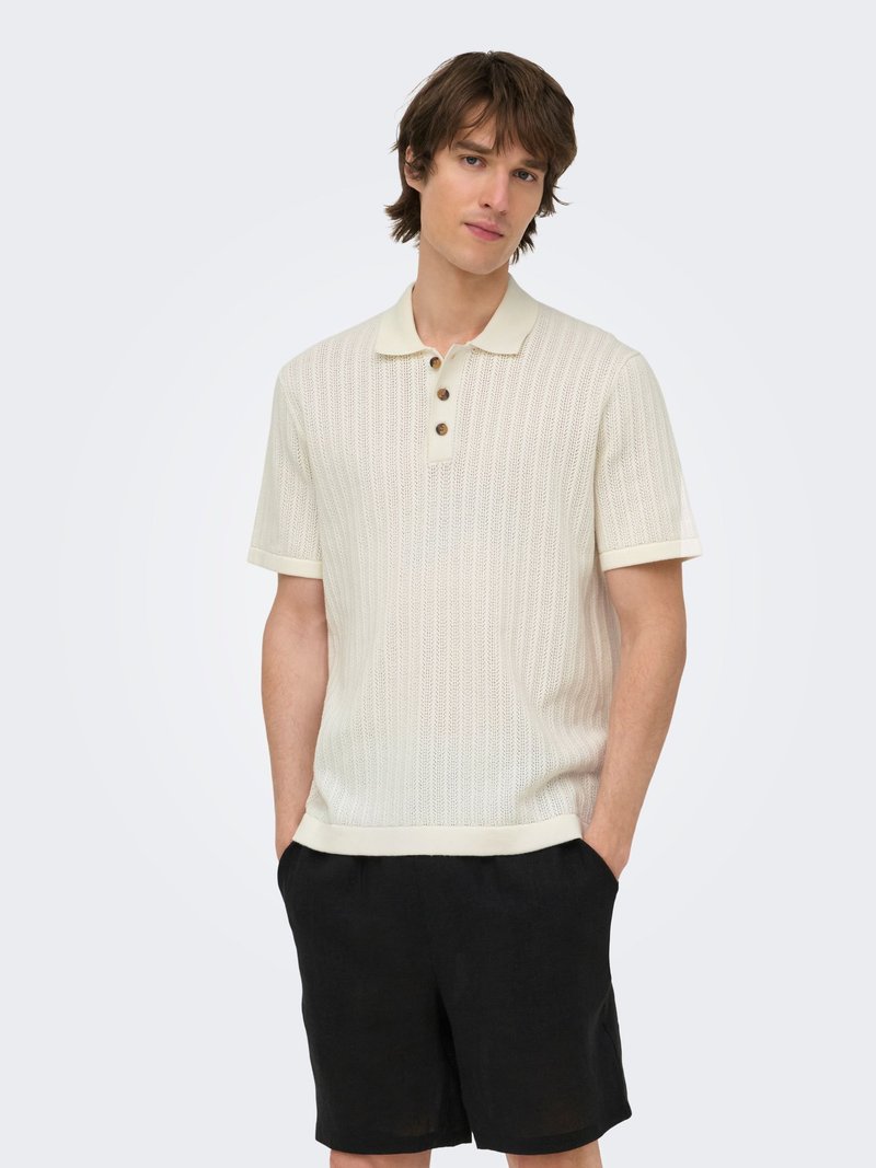 Jeune homme aux cheveux bruns portant un polo en maille crème à manches courtes et un short noir, debout les mains dans les poches sur fond blanc.