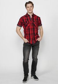 Camicia a maniche corte a quadri rossi e neri con due tasche sul petto, abbinata a jeans grigio scuro e sneaker nere, in piedi davanti a uno sfondo neutro.