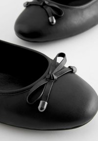 Ballerine nere realizzate in materiale sintetico liscio. Punta arrotondata, fiocco nero in raso con dettagli in plastica, design semplice e texture morbida.