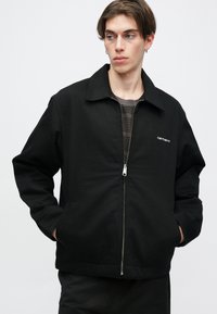 Μαύρο μπουφάν με μπροστινό φερμουάρ, επίπεδο γιακά και πλαϊνές τσέπες. Φτιαγμένο από υφασμάτινο με υφή, με μικρό λογότυπο στο στήθος.