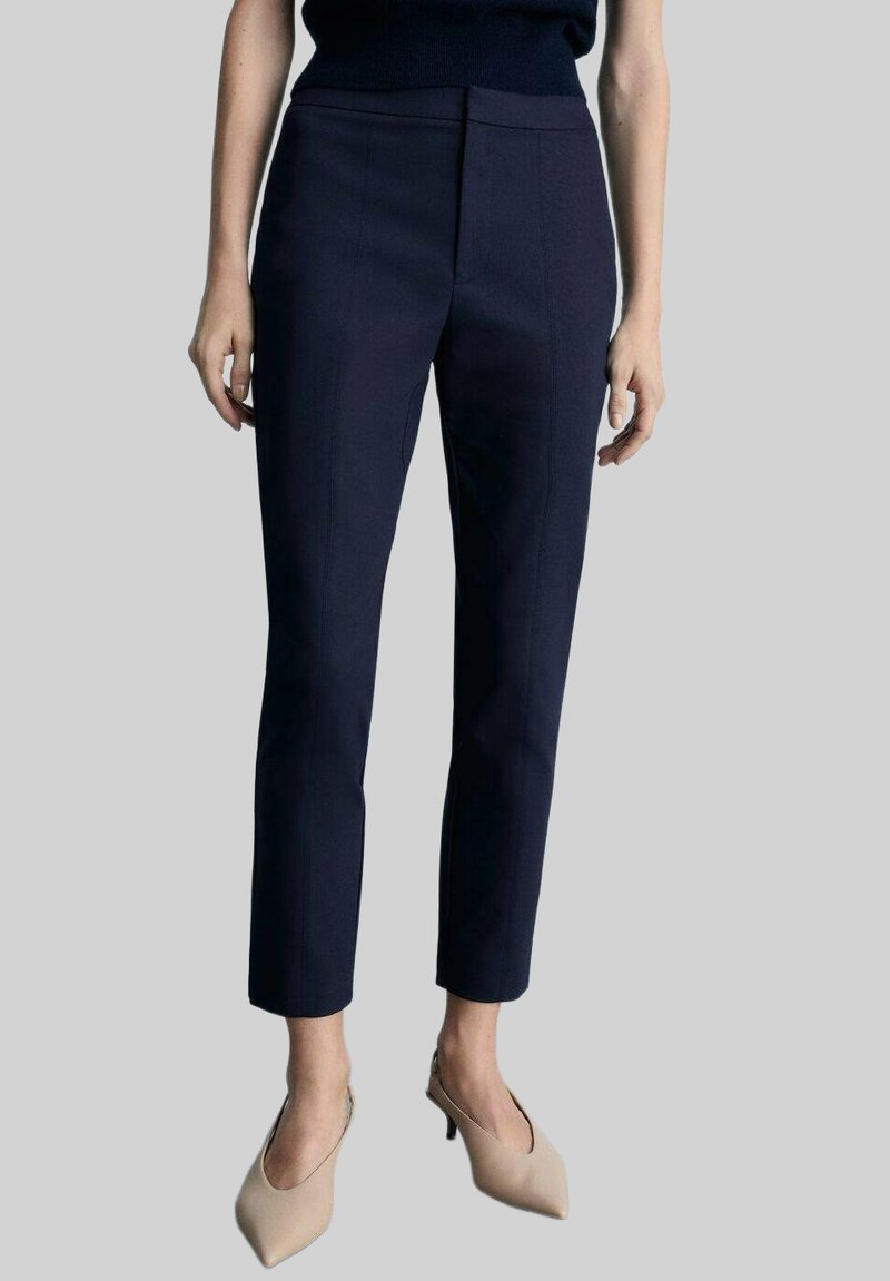 Navy-farbene, taillierte Hose mit slim fit, die eine glatte Textur und einen dezenten frontalen Bügelfalte aufweist. Kombiniert mit hellbeigen Stöckelschuhen.