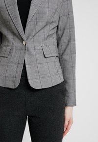 Blazer grigio a quadri con un design aderente, due tasche frontali e chiusura a un bottone. Abbinato a pantaloni grigio scuro su misura.