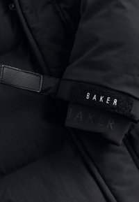 Detalj av en svart puffjacka med strukturerad tyg, elastiska ärmslut och en märkesvelcrorem med texten "BAKER." Slät och matt yta.