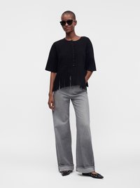 Femme portant un haut noir à franges boutonné, un jean gris à jambes larges avec revers, des chaussures noires à bout pointu cloutées, et des lunettes de soleil foncées sur un fond uni.