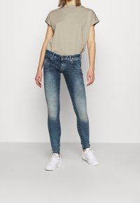 Mujer con una blusa beige de manga corta, jeans ajustados azules deslavados y zapatillas blancas de pie contra un fondo blanco liso.