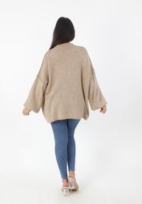 Beige oversized gebreide trui met wijde mouwen, een ribgebreide halslijn en een gebogen zoom, gecombineerd met blauwe skinny jeans en doorschijnende sandalen met hak.