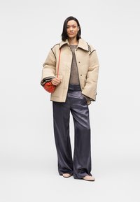 Γυναίκα που φοράει μπεζ μπουφάν με καπιτονέ λεπτομέρειες, γκρι σατέν παντελόνι φαρδιάς γραμμής, ανοιχτό καφέ πουλόβερ, μπεζ μπαλαρίνες και κρατάει μια μικρή κόκκινη τσάντα χειρός.