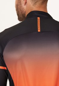 Svart och orange sporttröja med en gradientdesign, har en hög krage och en reflekterande logotyp nära bakre nacklinjen.