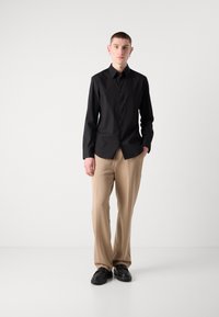 Camicia nera con bottoni e maniche lunghe, abbinata a pantaloni beige a gamba larga e scarpe chunky nere, con un tessuto liscio e una vestibilità su misura.