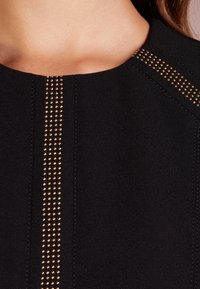 Musta kangastoppi, jossa on pyöreä pääntie ja pystysuorat rivit pieniä kultaisia niittejä harteilla, sileä pinta ja siisti ompelu.